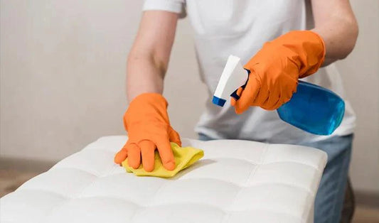 Person cleaning white mattress with spray and cloth, wearing orange gloves